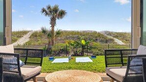 A balcony with wicker chairs, a woven mat, and a view of a grassy area with a palm tree and a fence. - Oceanfront Condo:1st Floor, Steps to the Ocean (Jacksonville Beach)