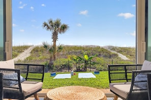A balcony with wicker chairs, a woven mat, and a view of a grassy area with a palm tree and a fence.