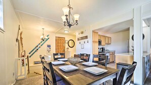 Un comedor con una mesa de madera dispuesta para cuatro personas, rodeado de sillas. Un candelabro cuelga arriba, y esquíes están colgados en la pared. Se ve una cocina al fondo.