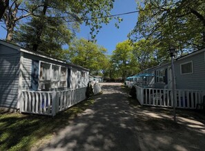 A row of mobile homes with white railings, a paved driveway, and trees in the background.