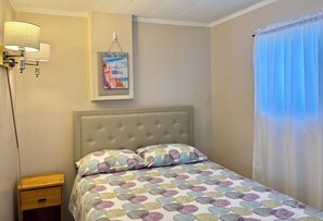 A bedroom with a tufted headboard, a bed with a polka dot bedspread, a nightstand with a drawer, a wall-mounted lamp, and a beach-themed poster.