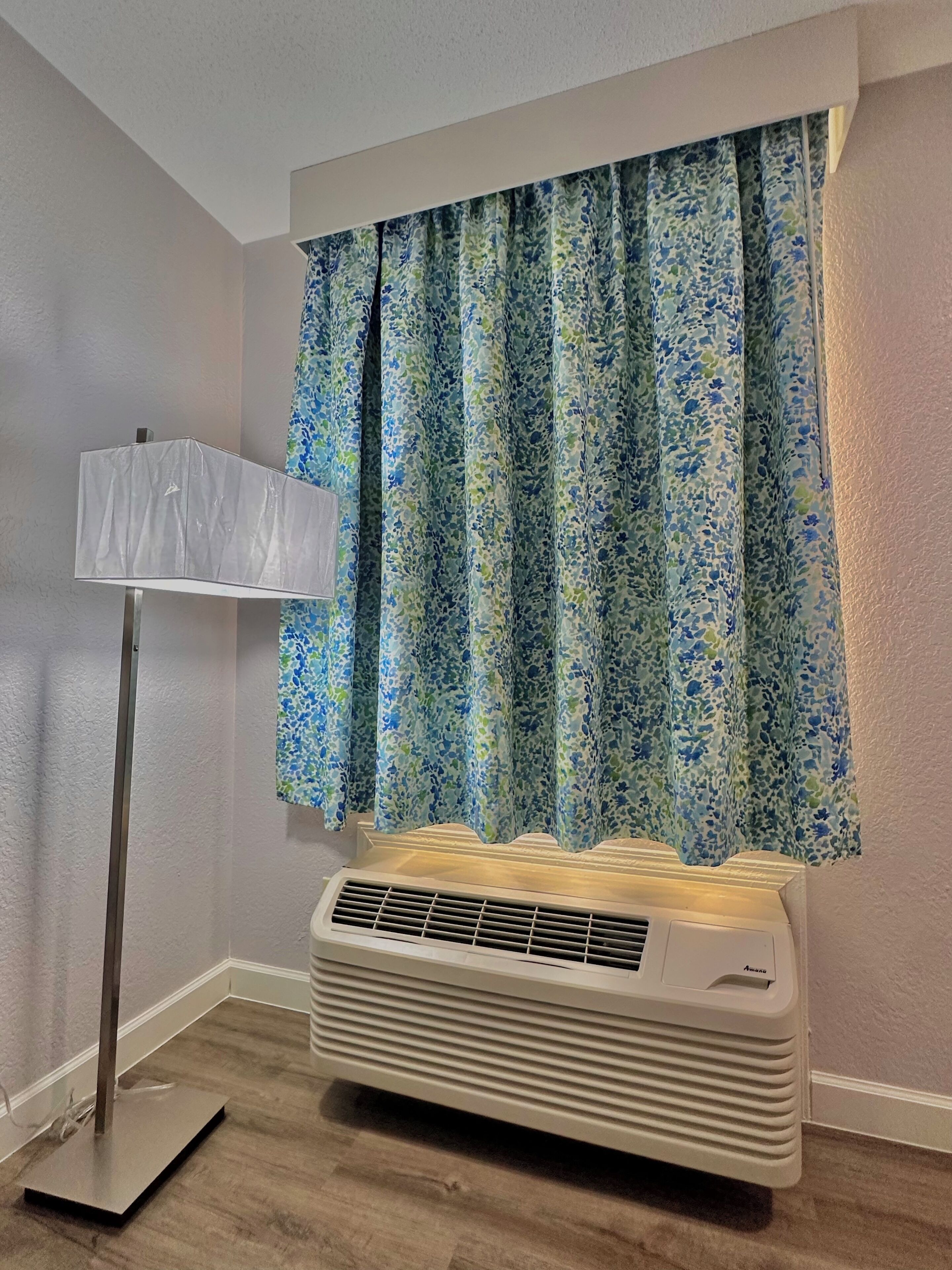 Una habitación con una ventana con cortinas florales, una lámpara de pie y un aire acondicionado blanco.