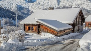 Een houten hut met een met sneeuw bedekte dak, omgeven door een besneeerd landschap en bergen op de achtergrond.
