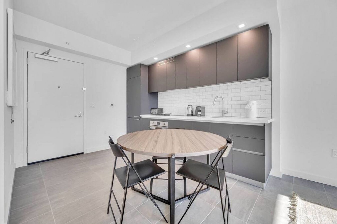 A modern kitchen with a round wooden table and black chairs, a white door, and grey cabinets.