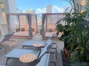 A rooftop lounge with sun loungers, small round tables, and potted plants.