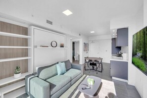 Ein modernes Wohnzimmer mit einem grauen Sofa, einem Couchtisch und einem Essbereich mit Tisch und Stühlen. Weiße Wände, ein Bücherregal und ein Gemälde eines Waldes sind zu sehen.