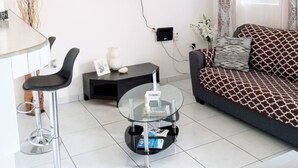 A modern living room with a flat-screen TV mounted on the wall, a round glass coffee table, a sofa with patterned cushions, and a dining area with a white table and two bar stools. - Cozy, Cole Bay, one bedroom with security and free parking (Cole Bay)