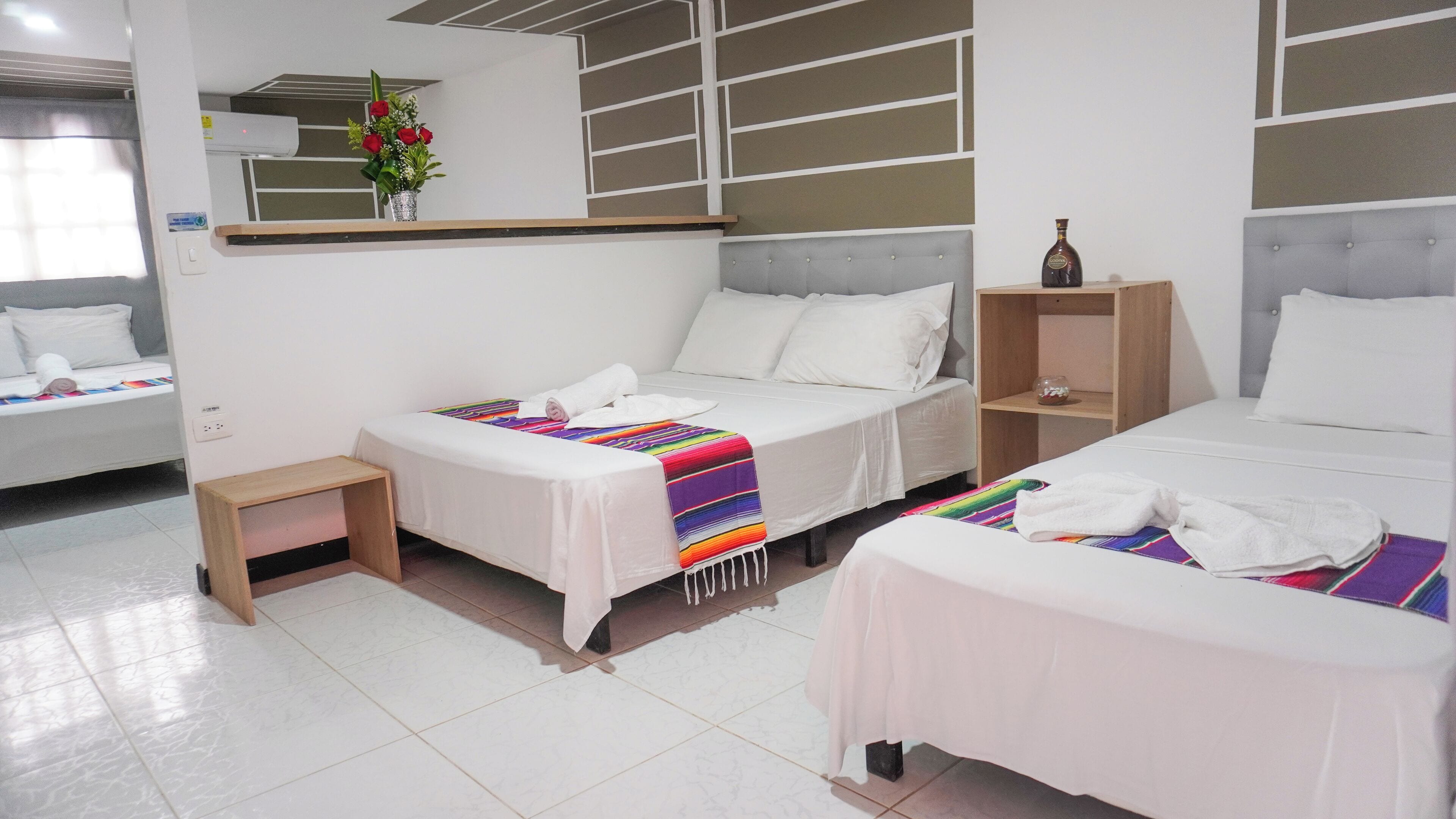 Une chambre d’hôtel avec trois lits, chacun recouvert de draps blancs et d’une couverture rayée aux couleurs vives. On y trouve une petite table de chevet en bois sur laquelle est posé un récipient en verre, ainsi qu’une étagère fixée au mur, sur laquelle se trouve une bouteille.