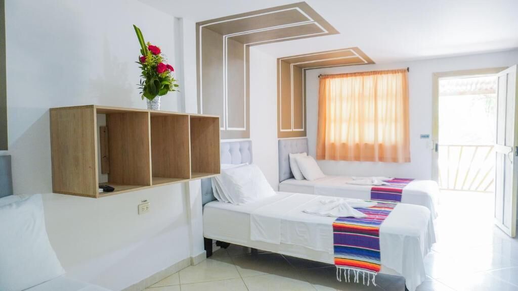 A hotel room with two beds, a wooden shelf, a vase with flowers, and a window with curtains.