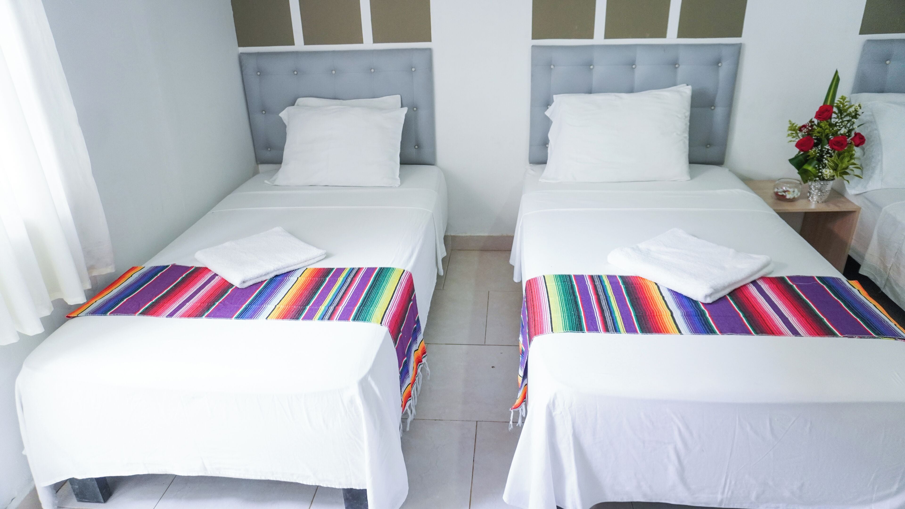 Two single beds with white linens and colorful striped bedspreads, a grey upholstered headboard, and a small table with a vase of red flowers.