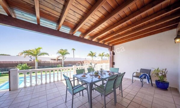 Une terrasse couverte avec une table dressée pour les repas, des chaises, une plante en pot et une vue sur une piscine et des palmiers.