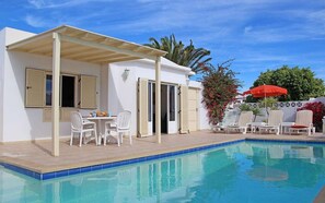 Un espace piscine doté d’un patio couvert, d’une table avec des chaises et d’un parasol rouge.