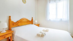 Een net opgemaakt bed met witte lakens en opgerolde handdoeken, een houten hoofdbord, een bedlampje en een raam met dunne gordijnen.