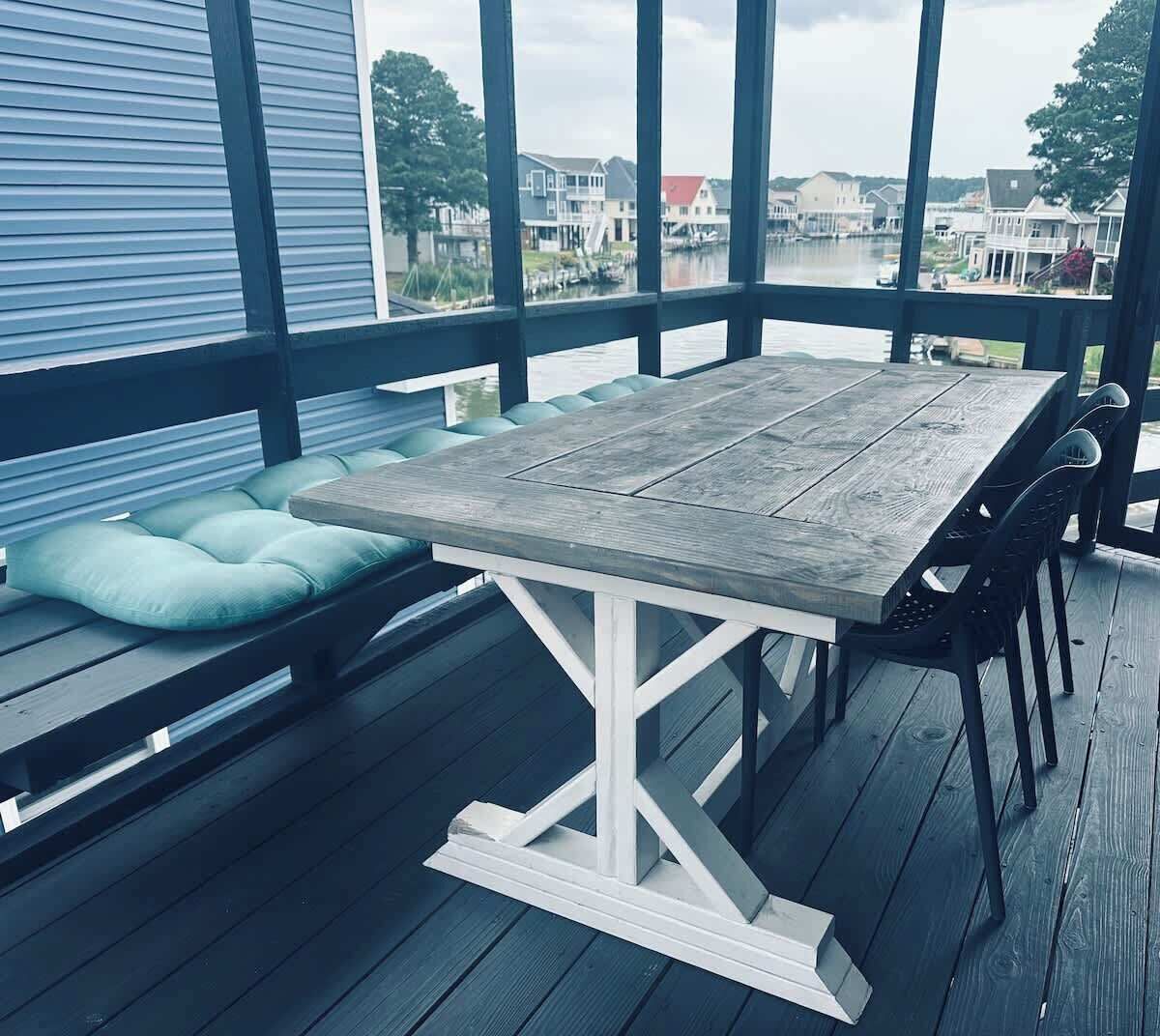 A wooden deck with a table and chairs, a cushioned bench, and a view of houses across a body of water.