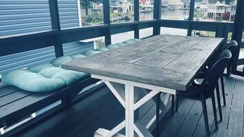 A wooden deck with a table and chairs, a cushioned bench, and a view of houses across a body of water.