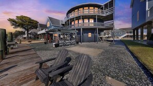 Una propiedad frente al agua con muelle de madera, mobiliario exterior y un edificio de varios niveles con balcones.