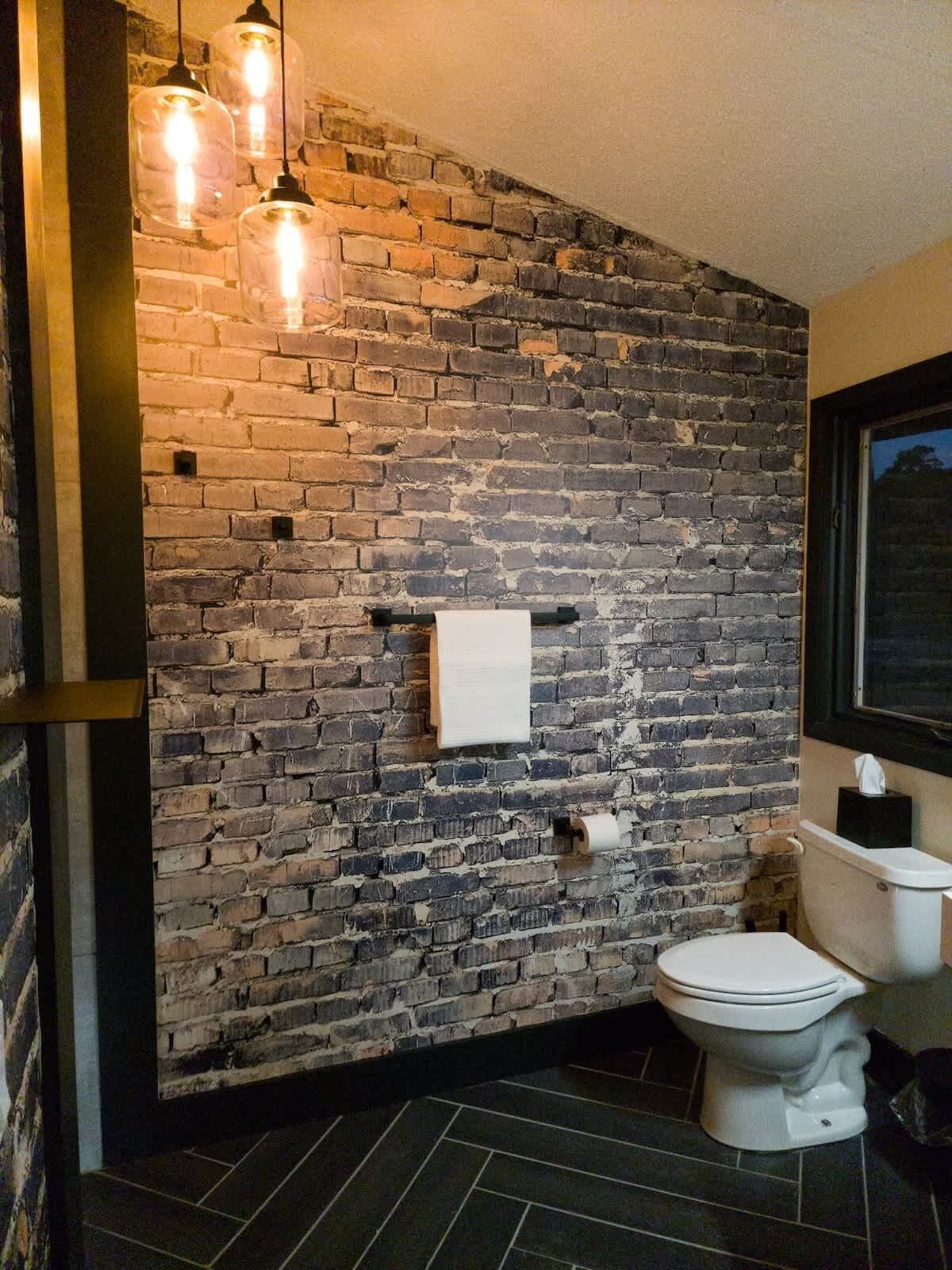 A bathroom with a brick wall, a toilet, and hanging pendant lights.