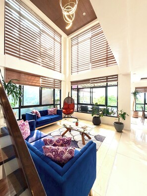 A spacious living area with a blue sofa set, a glass coffee table, and a hanging chair. Large windows allow natural light and views of the outdoors. - Picturesque private villa with infinity pool (Puerto Galera)