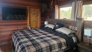 A wooden cabin room with a plaid bedspread, a television mounted on the wall, and a window with curtains. - Scenic Getaway - Rustic Cabin - Mountain Ridge (Pinon Hills)