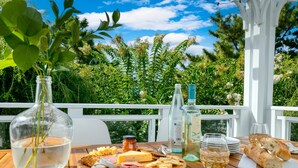 Ein Holztisch mit einer Wurstplatte, einer Wasserflasche und einem Weinglas auf einer Terrasse im Freien, mit weißer Reling und Grünpflanzen im Hintergrund.