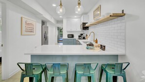 Una cocina moderna con armarios blancos, una isla central con taburetes y lámparas colgantes.