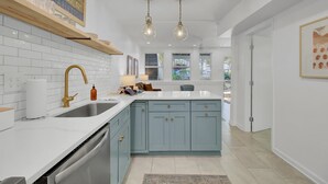 Una cocina moderna con azulejos blancos tipo metro, un lavamanos de acero inoxidable y una isla azul. Se observan luces colgantes tipo lámpara y un estante de madera sobre el lavamanos.