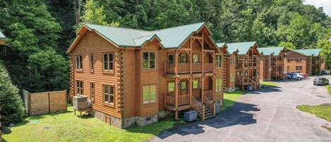 A row of log cabins with multiple stories and balconies, surrounded by a forested area.