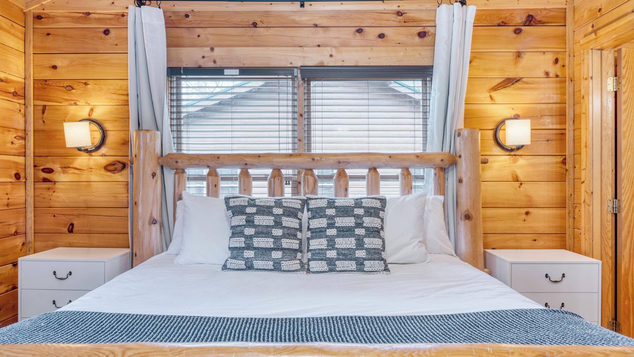 Une chambre à coucher de style cabane en bois, avec un lit, deux tables de chevet, une fenêtre avec stores et des lampes fixées au mur.