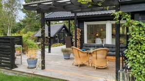 A wooden deck with wicker furniture, a pergola, and a view of a cabin with large windows. - Beautiful home in Vig with WiFi (Vig)