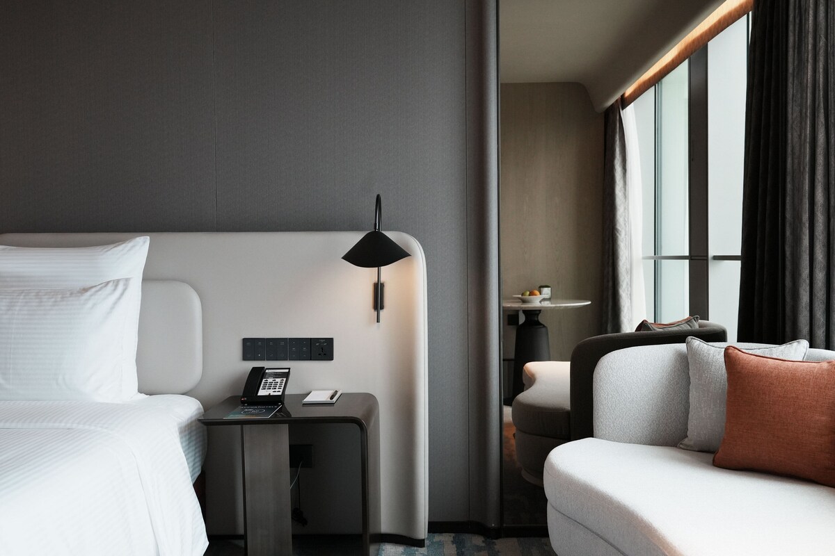 A modern hotel room with a bed, a desk with a phone, a lamp, a sofa, and a window with curtains.