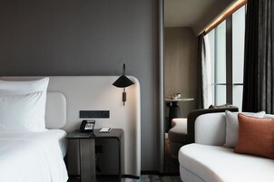 Ett modernt hotellrum med en säng, ett skrivbord med en telefon, en lampa, en soffa och ett fönster med gardiner.