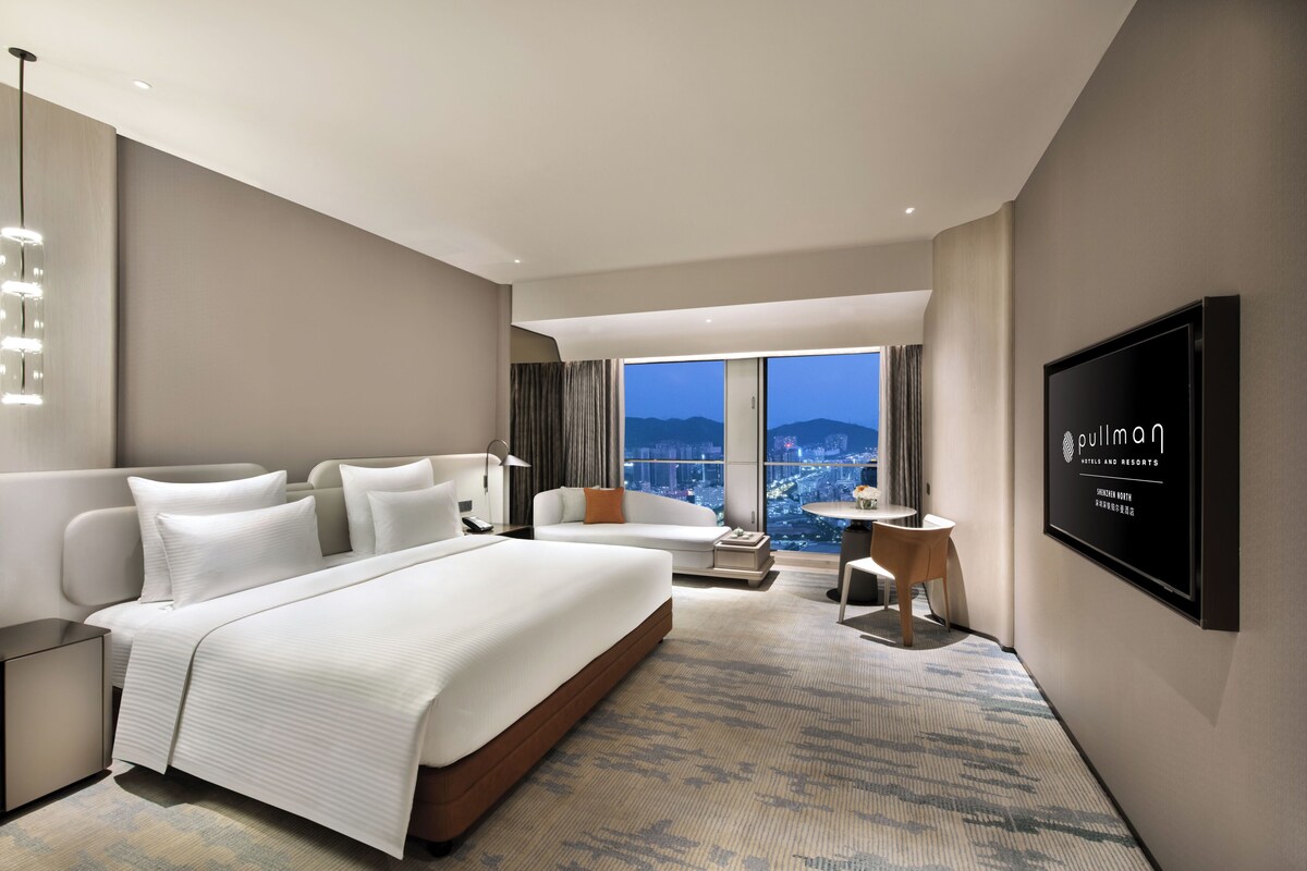 A modern hotel room with a large bed, a sofa, a TV, and a city view.