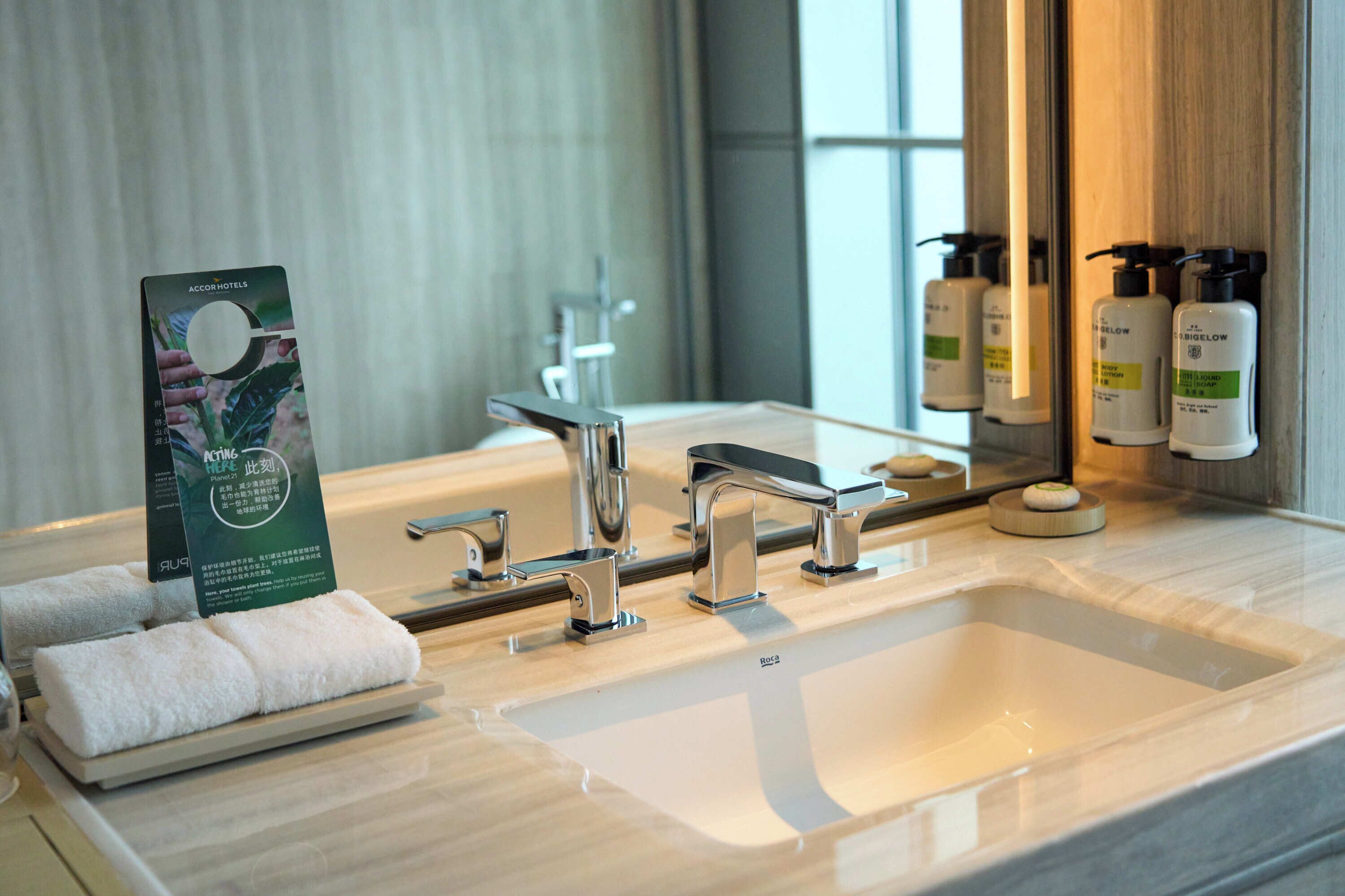 A hotel bathroom with a large sink, two faucets, and a mirror. There are towels on a tray and toiletries on the counter.