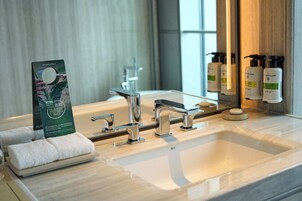 A hotel bathroom with a large sink, two faucets, and a mirror. There are towels on a tray and toiletries on the counter.