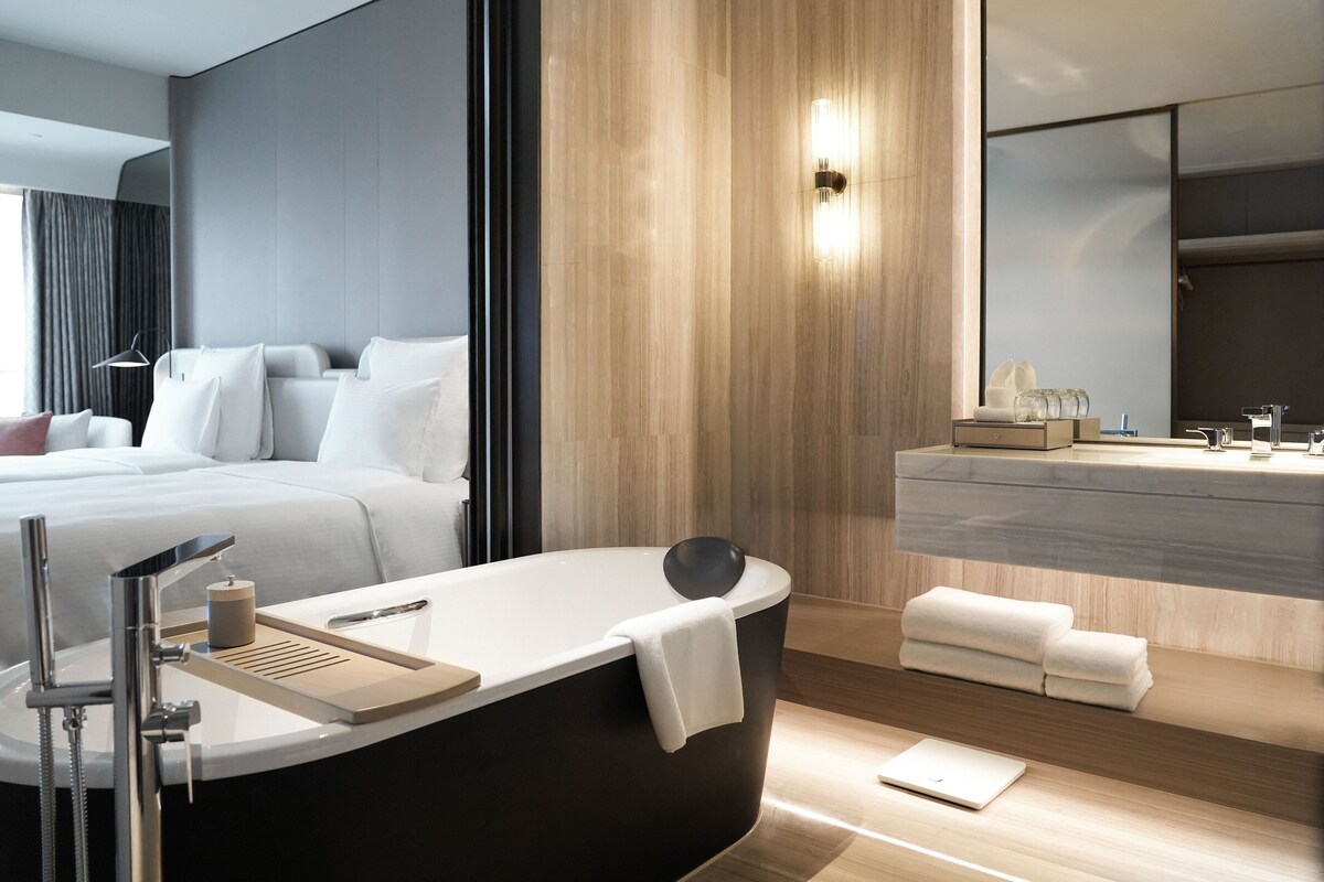 A modern hotel room with a large bed, a bathtub, a vanity with a mirror, and a wooden wall.