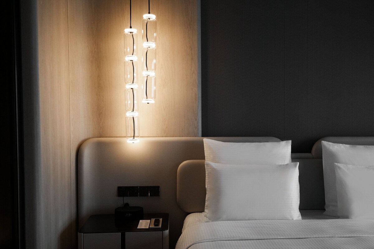 A modern hotel room with a bed, bedside table, and a wall-mounted light fixture.