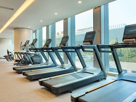 Une rangée de tapis de course dans une salle de sport dotée de grandes fenêtres et offrant une vue sur un immeuble.