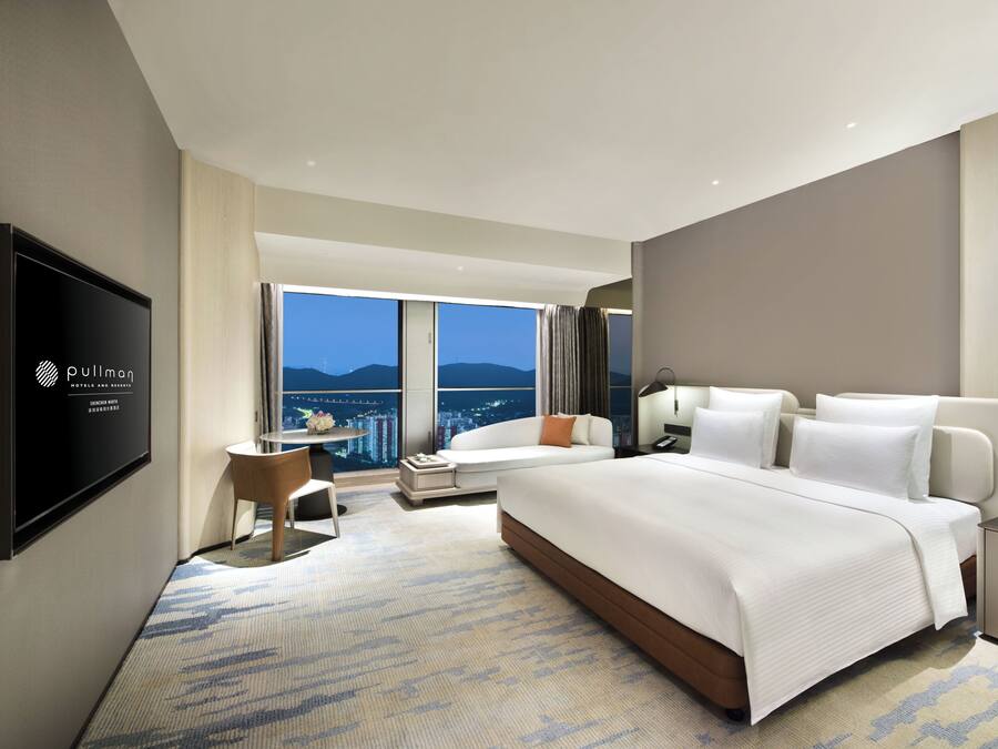 A modern hotel room with a large bed, a desk, a chair, a TV, and a view of the cityscape.