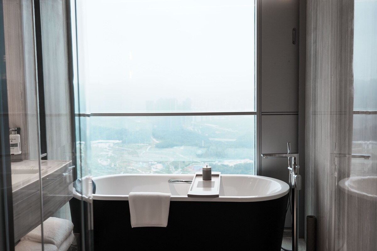 A modern bathroom with a freestanding bathtub, a large window offering a city view, and a glass shower enclosure.