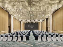 A conference room with rows of tables and chairs, a large screen at the front, and a ceiling with numerous lights.