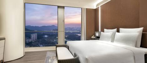 A modern hotel room with a large bed, a seating area, and a city view through large windows.