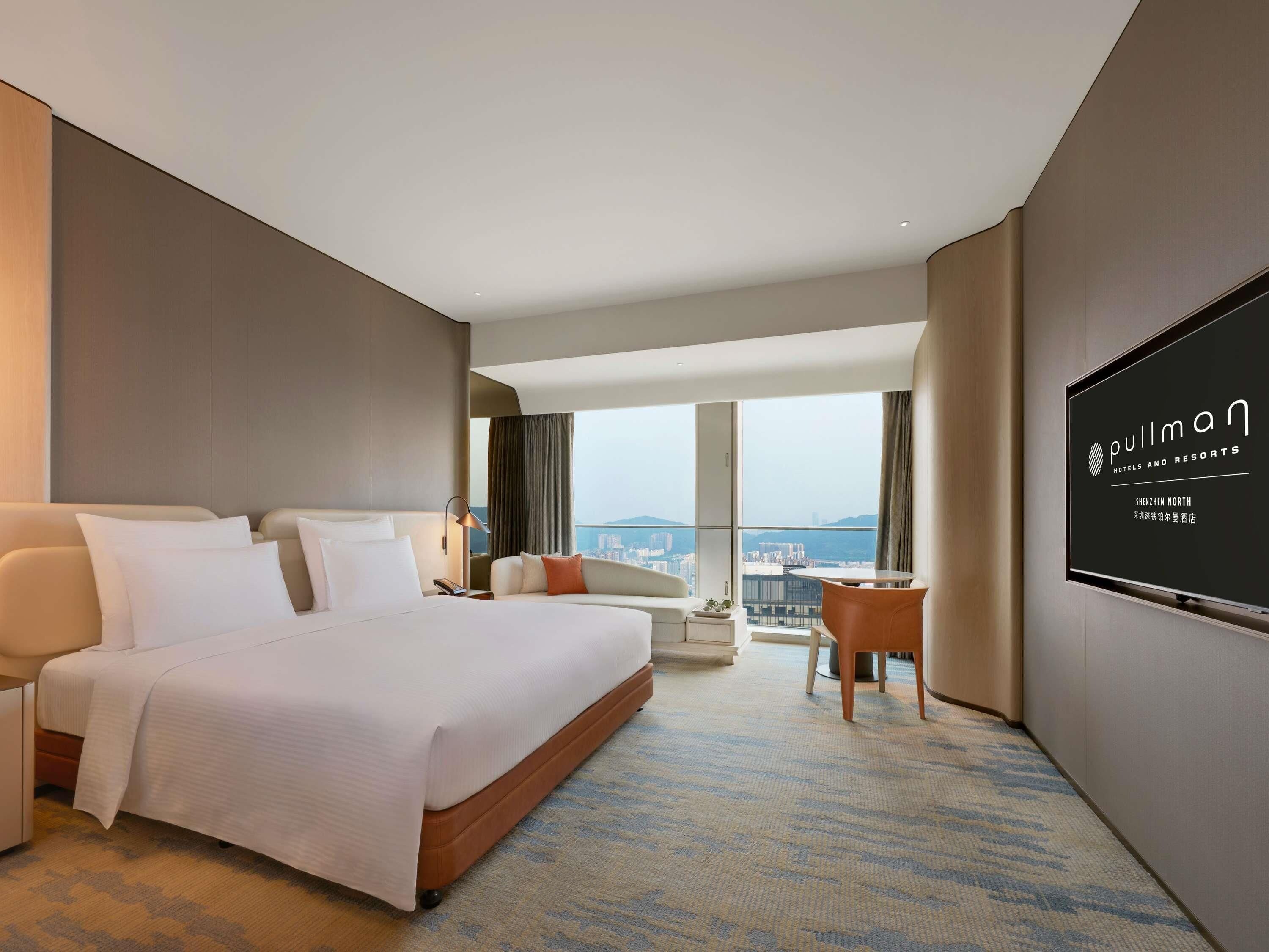 A hotel room with a large bed, a desk, a chair, a TV, and a view of the city.
