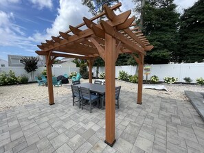 A wooden pergola with a table and chairs underneath, surrounded by a paved patio area.