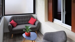 Een balkon met een grijze bank, een rode kussen, een kleine tafel met een plantje en een grijze fauteuil.