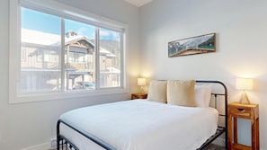 Une chambre avec une grande fenêtre, un lit recouvert de linge de lit blanc, deux tables de chevet avec des lampes et un tableau encadré au mur.