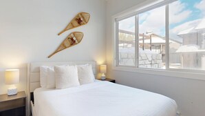 Un dormitorio con cama blanca, dos elementos decorativos en la pared, una mesita de noche con lámpara y una ventana con vistas a edificios y nieve.