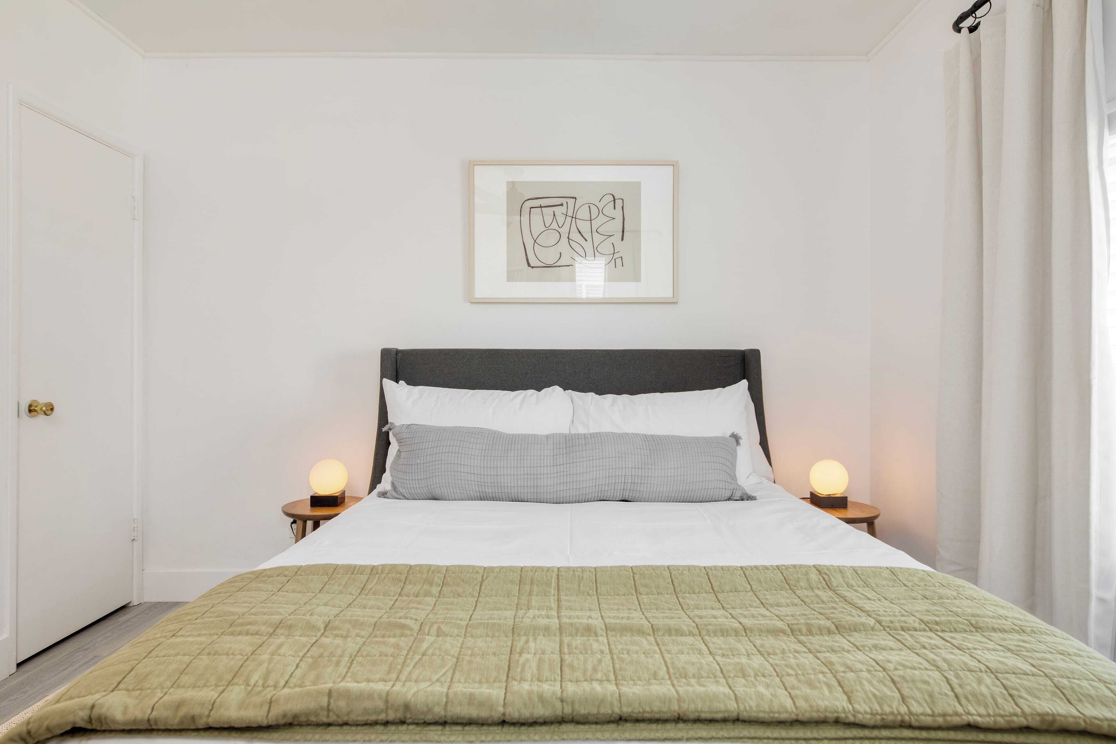 Un lit bien fait, recouvert d’une couette verte, avec des oreillers blancs et une œuvre d’art abstraque encadrée au-dessus. Deux lampes de chevet sont placées de chaque côté du lit.