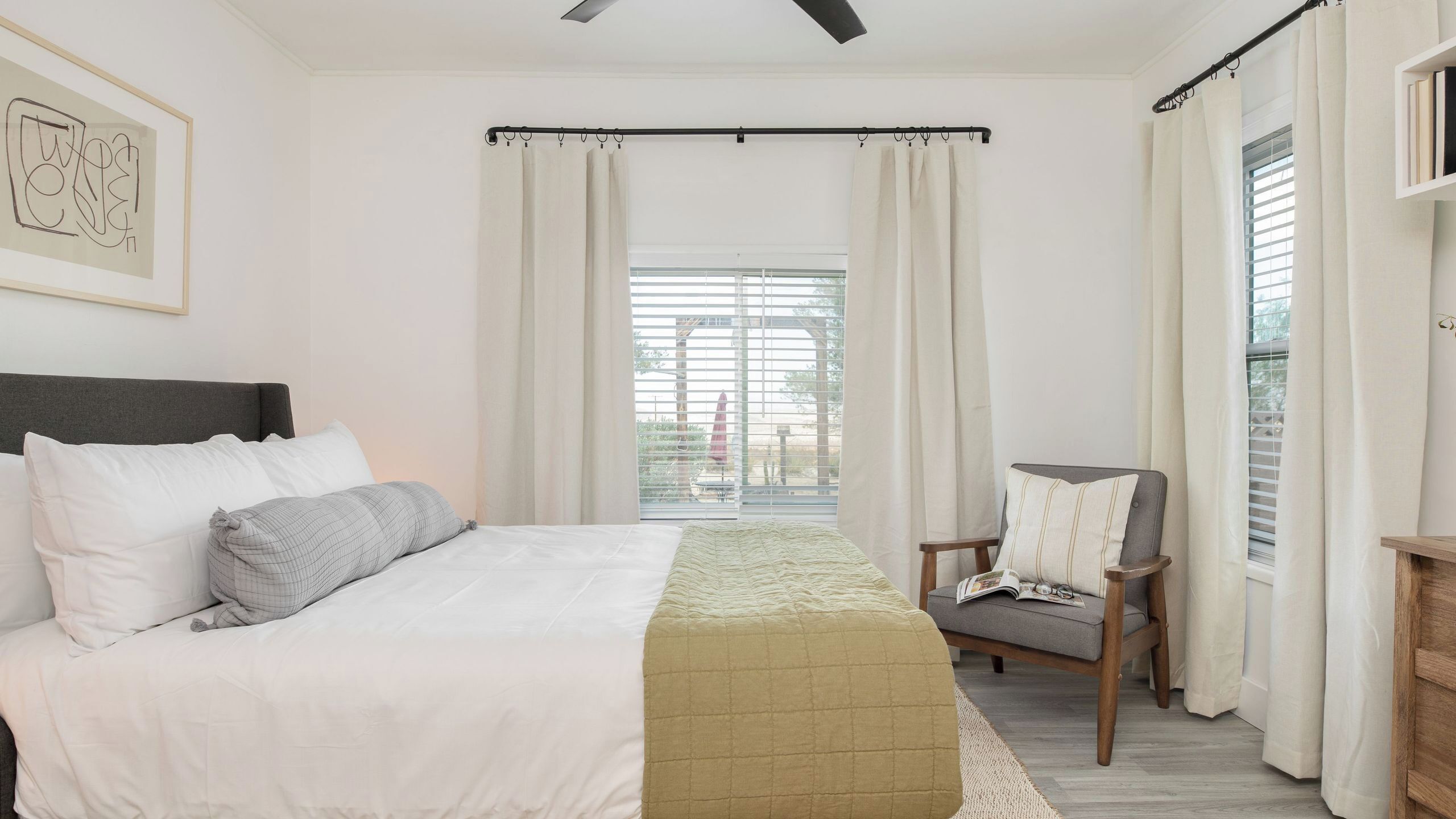 A bedroom with a bed, a chair, a window with curtains, and a ceiling fan.