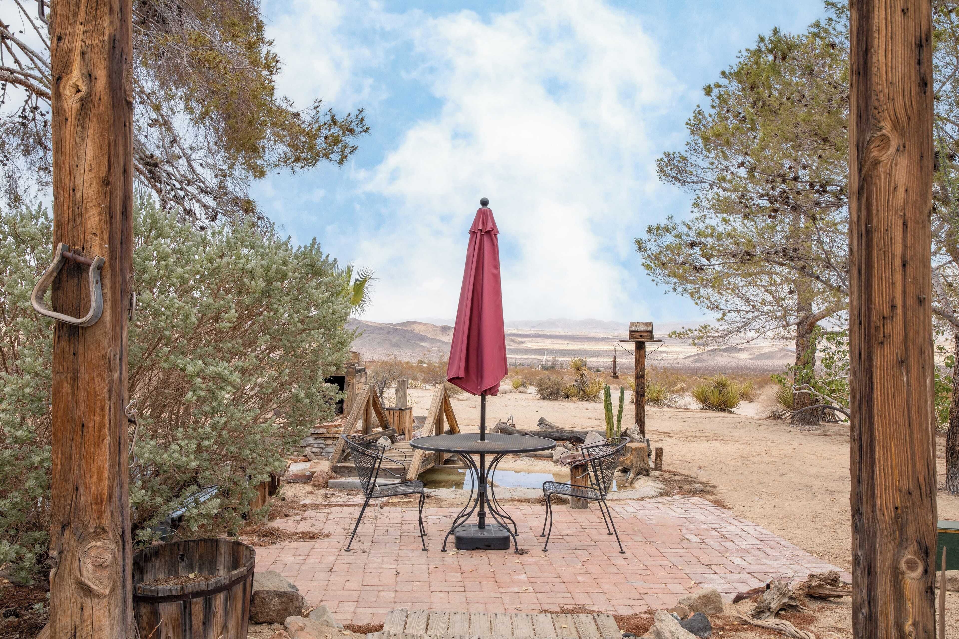 Een terras buiten met een tafel en stoelen, omringd door houten palen en een rode parasol.