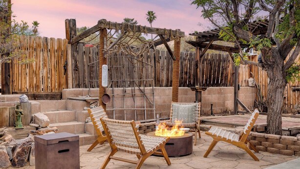 An outdoor patio with two wicker chairs, a round table with a fire pit, and a wooden pergola. - Boho Joshua Tree Escape w/ Firepit, Courtyard & Epic Views Asteroid by Avantstay (Twentynine Palms)